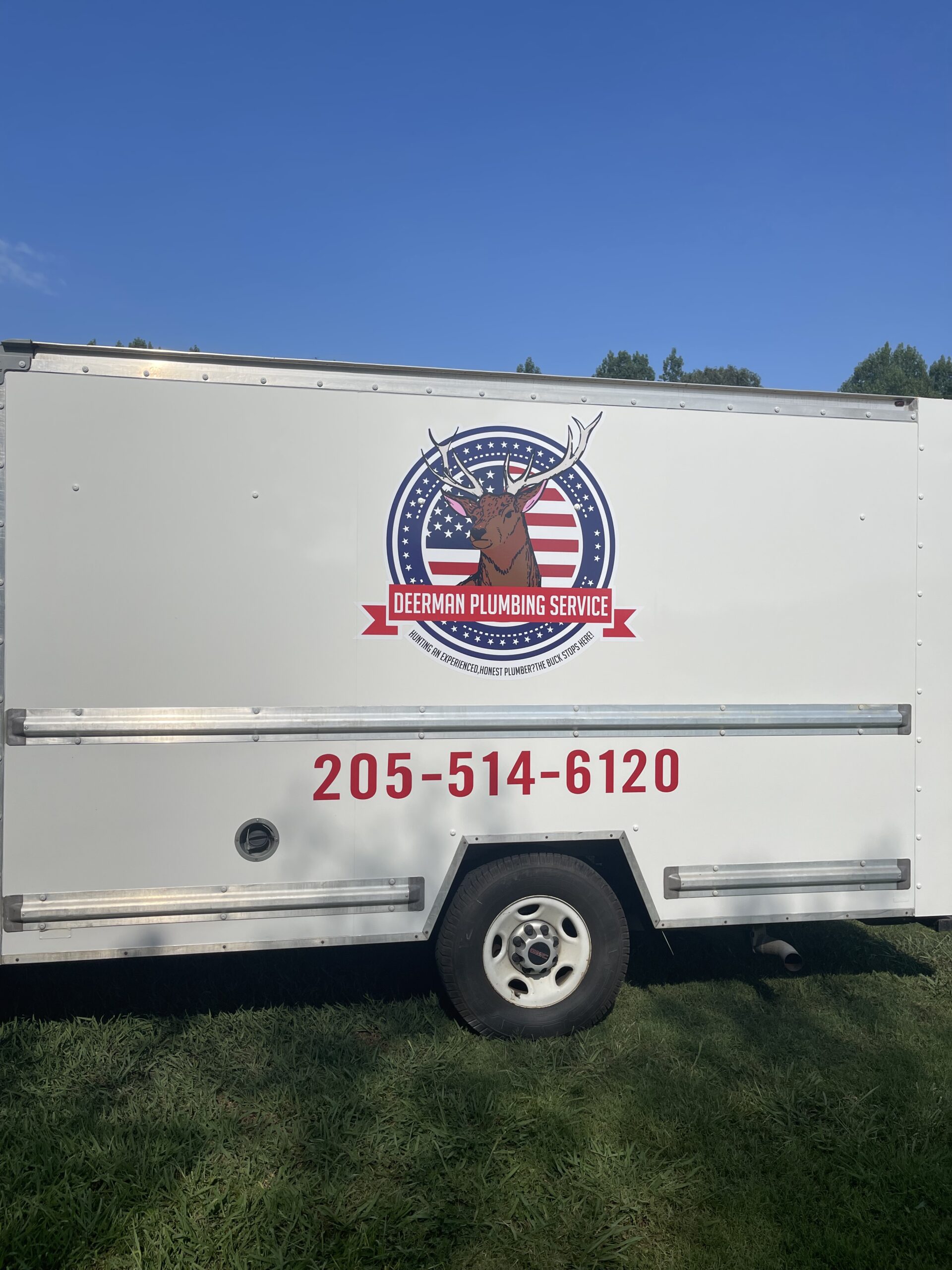 White work trailer with a Deerman Plumbing Service logo featuring a deer head and American flag. Red contact number 205-514-6120 is displayed below the logo.