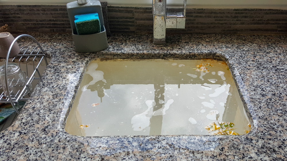 A kitchen sink filled with cloudy, dirty water, food debris, and soap suds, with a sponge and dish rack nearby on a granite countertop.