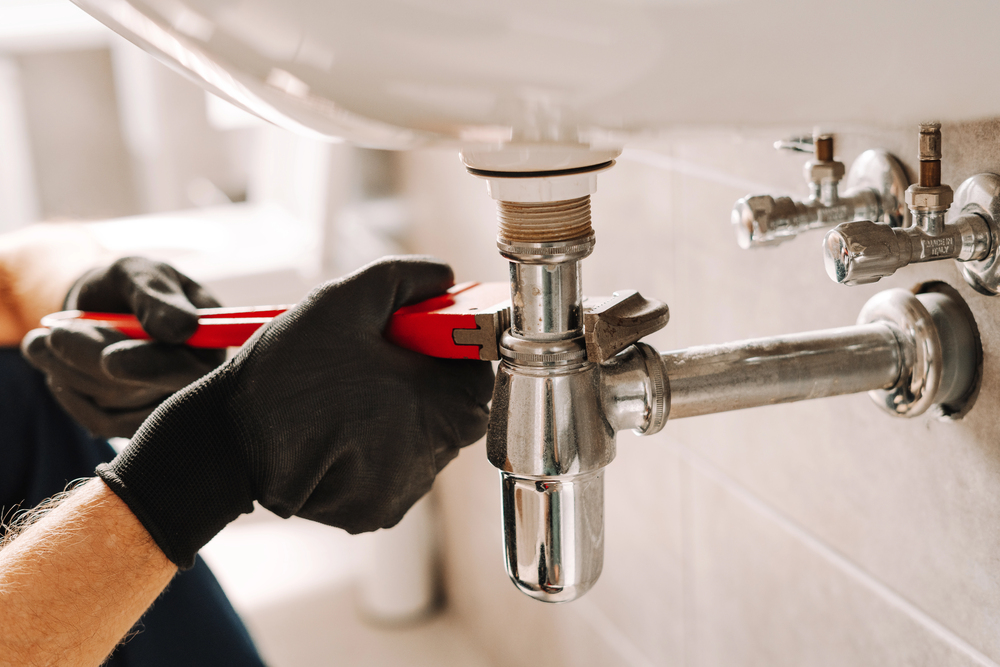 Person wearing black gloves uses a red adjustable wrench to tighten a chrome pipe under a sink in a bathroom.