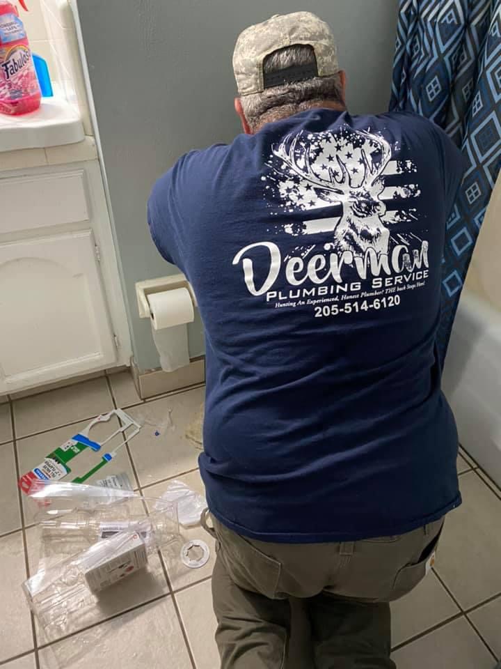 A plumber in a Deer Man Plumbing Service shirt installs or repairs a toilet paper holder in a bathroom with scattered tools and packaging on the tile floor.
