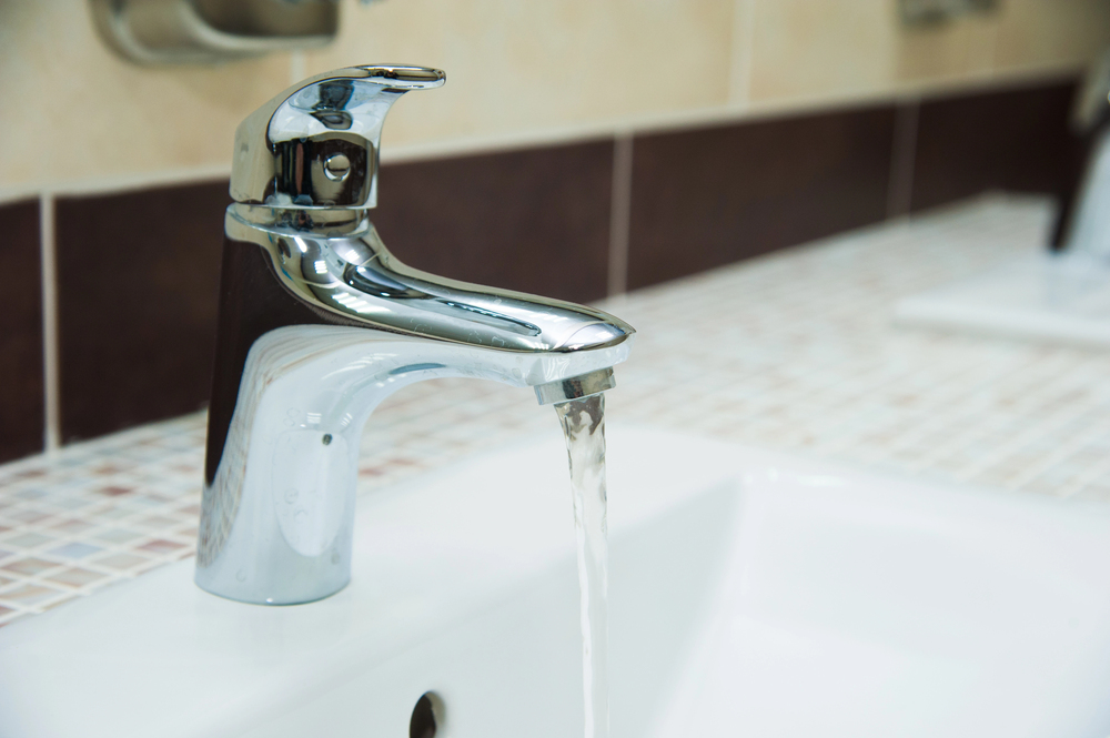 A chrome bathroom faucet with water running into a white sink, set against a tiled countertop and wall.