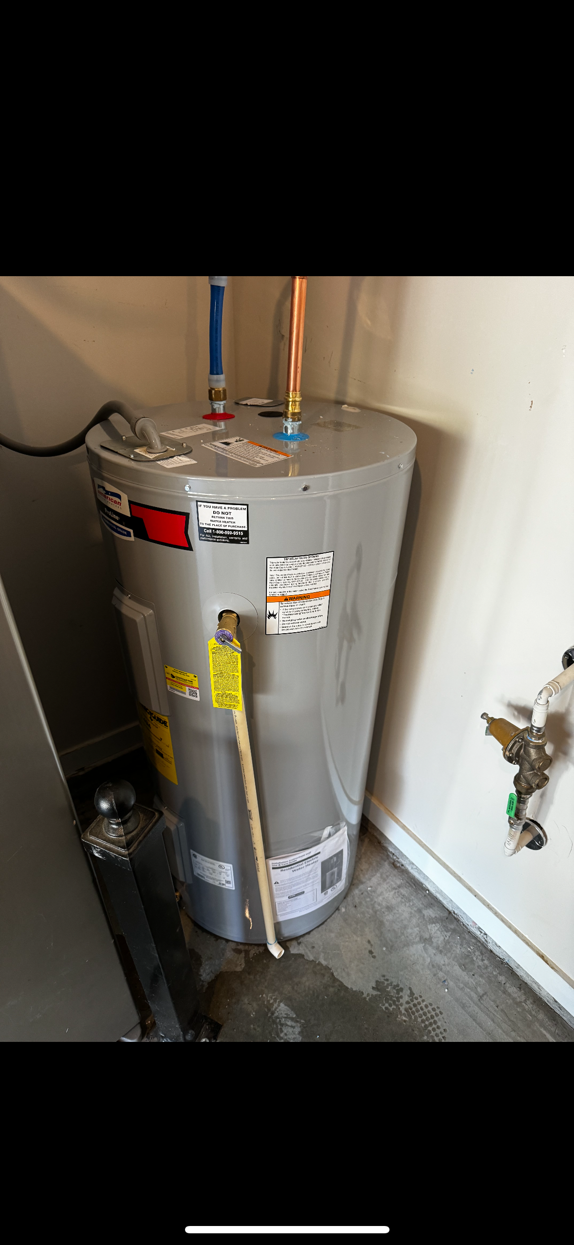 A gray water heater with attached pipes and warning labels is installed in the corner of a room with concrete flooring.