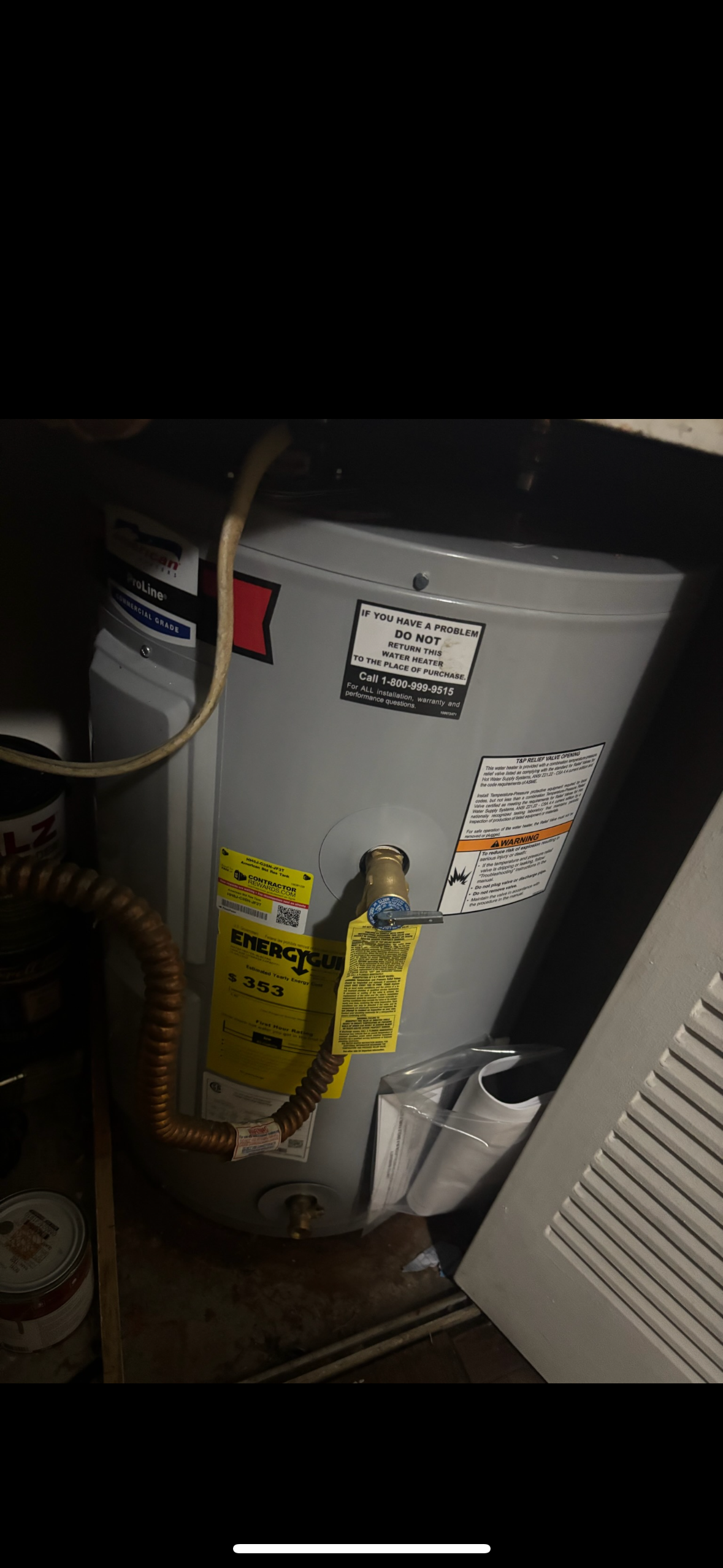 A gray water heater with multiple warning and information labels is installed in a small utility closet, surrounded by pipes and other household items.