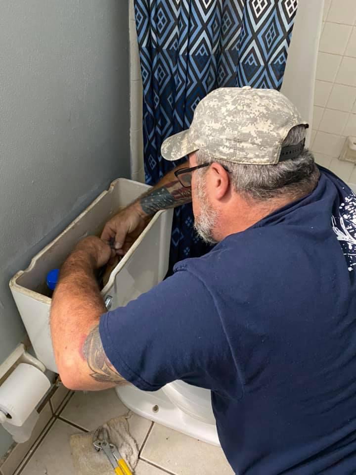 A man in a cap and glasses repairs the inside of a toilet tank in a bathroom with a blue and black patterned shower curtain.