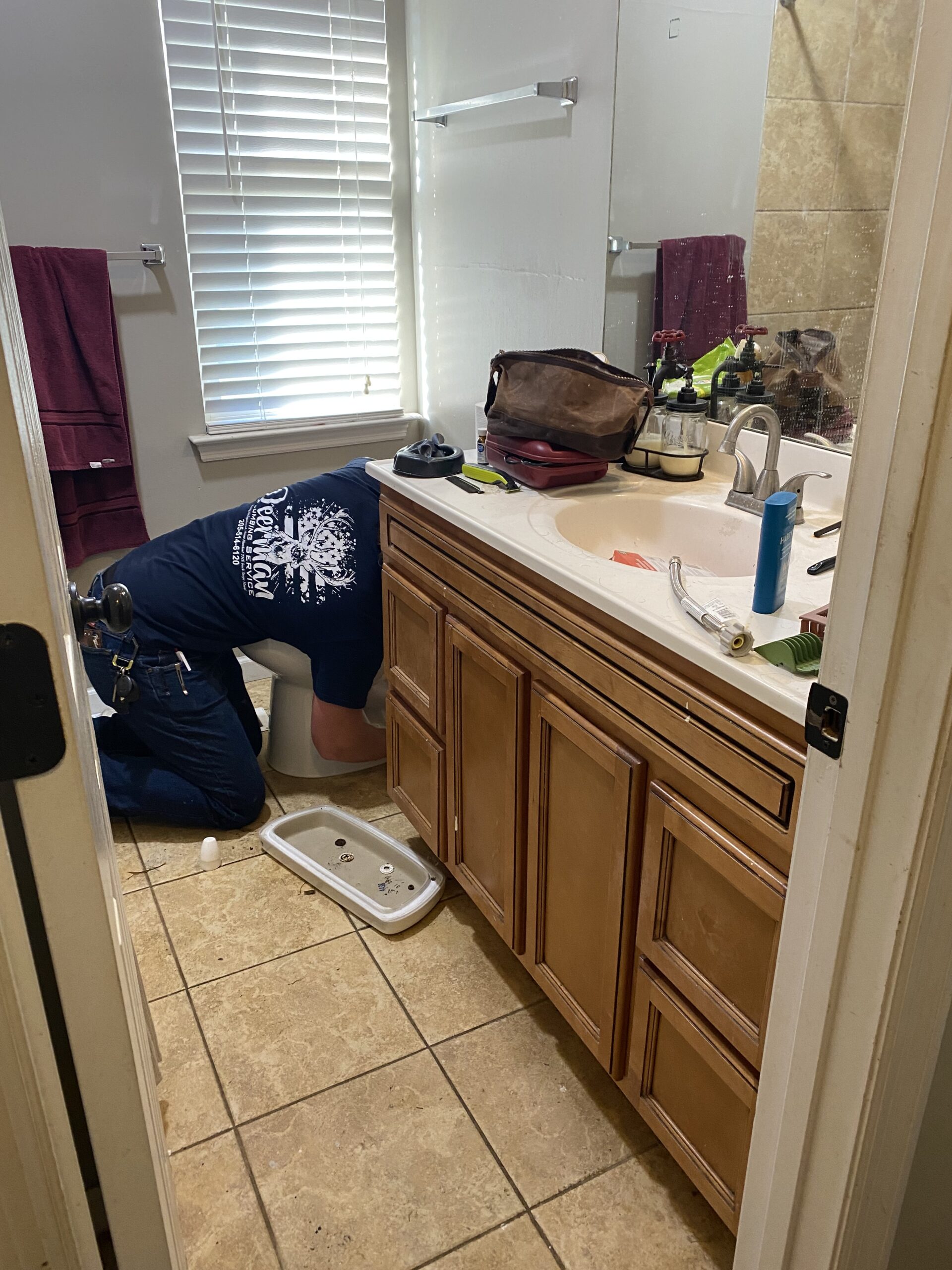 A person kneels in front of a toilet in a bathroom, appearing to perform maintenance or repair work. Tools and personal items are visible on the sink counter.