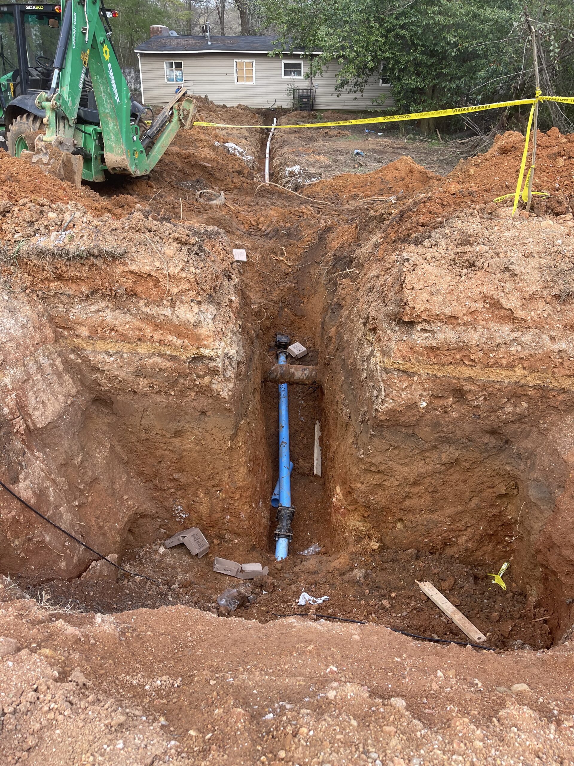 A trench dug in the ground exposes a blue pipe, with a backhoe and caution tape nearby; a house and trees are visible in the background.